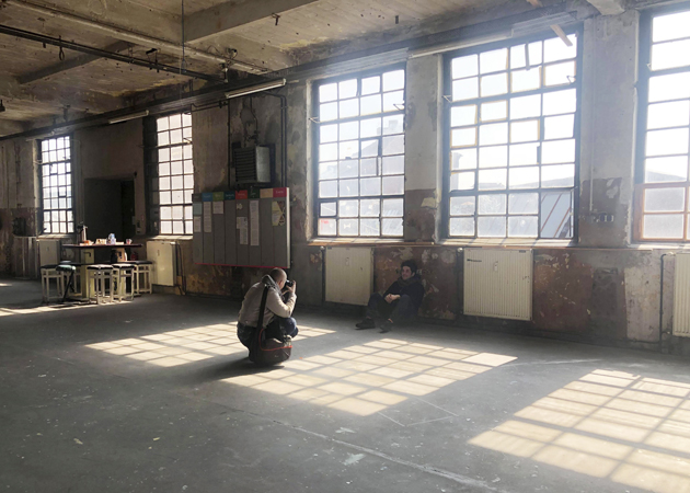 En studerande sitter på huk och fotograferar en annan som sitter lutad mot väggen i en gammal fabriksbyggnad som nu fungerar som som arbetsrum för konstnärer. Ljuset kommer vackert in genom fönstren.