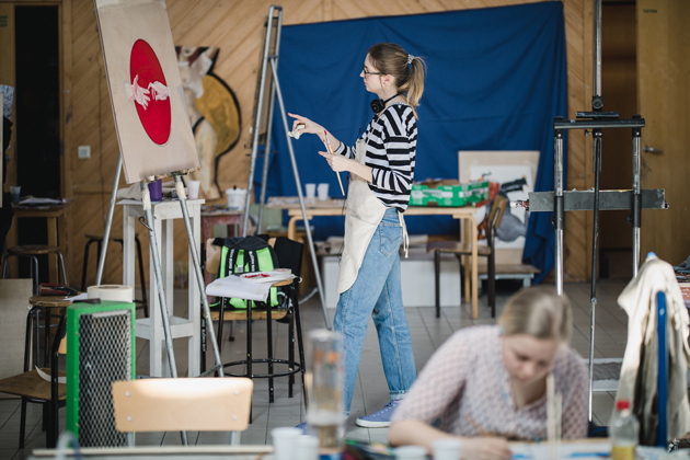 Bild från skolans ateljé där två studerande målar. Den ena sitter vid ett bord och den andra står vid ett staffli.